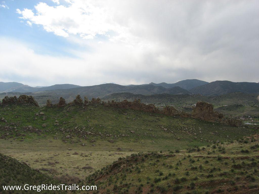 A panoramic view of rolling hills and rocky formations under a partly cloudy sky, showcasing vibrant green vegetation and distant mountains in the background. Devil's Backbone mountain bike trail.