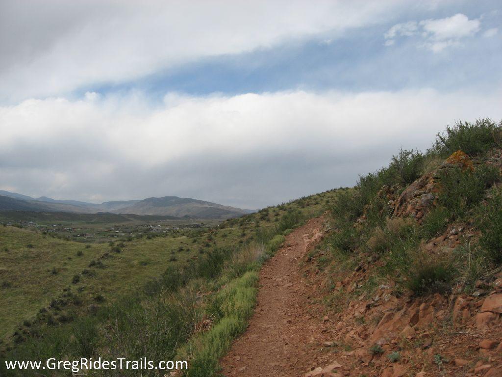 A winding dirt trail runs along a hillside, surrounded by lush green vegetation and rocky terrain. The scene features a cloudy sky overhead, with distant mountains visible in the background. The trail appears well-used and invites exploration of the natural landscape. Devil's Backbone mountain bike trail.