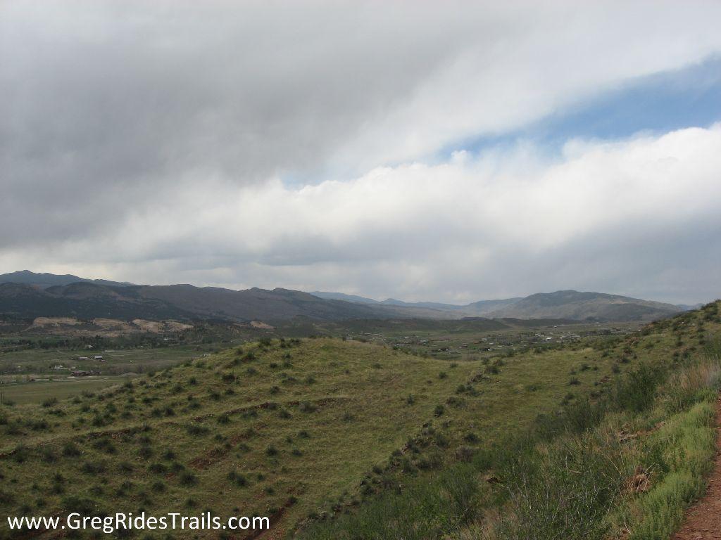 A panoramic view of rolling green hills under a cloudy sky, with distant mountains visible on the horizon. The landscape features patches of shrubs and grass, suggesting a natural, serene environment. Devil's Backbone mountain bike trail.