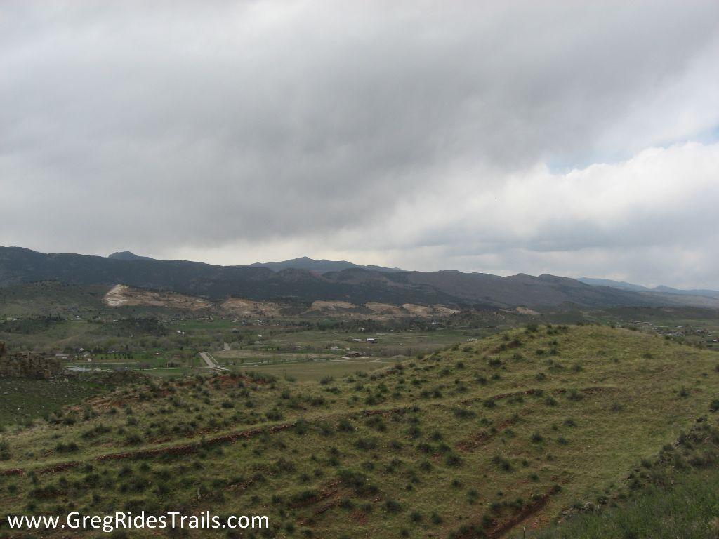 A scenic view of rolling hills and mountains under a cloudy sky, with patches of grass and small shrubs in the foreground. The landscape features a winding road and distant farmland, creating a peaceful and natural atmosphere. Devil's Backbone mountain bike trail.