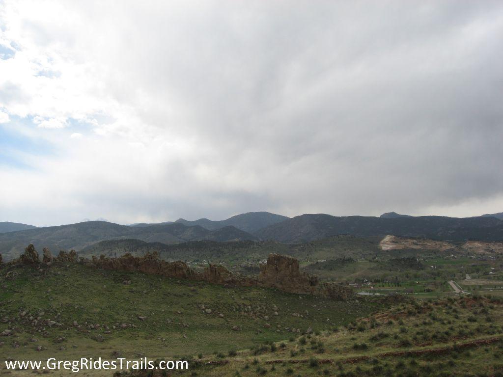 A scenic view of rolling hills and distant mountains under a cloudy sky. The landscape features rocky outcrops in the foreground, with patches of green grass and sparse vegetation. In the background, the mountains rise against the overcast sky, creating a tranquil natural setting. Devil's Backbone mountain bike trail.