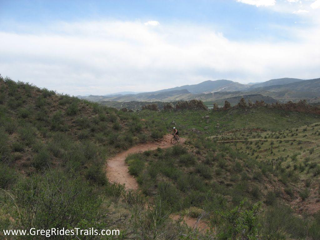 A mountain biker riding along a winding dirt trail in a hilly green landscape, with distant mountains and a cloudy sky in the background. Devil's Backbone mountain bike trail.