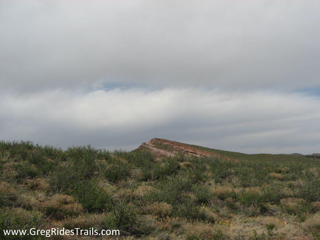 A wide landscape featuring rolling hills covered in green vegetation and sparse shrubs, under a cloudy sky. A rocky outcrop is visible in the background, contrasting with the soft, grassy terrain in the foreground. Devil's Backbone mountain bike trail.