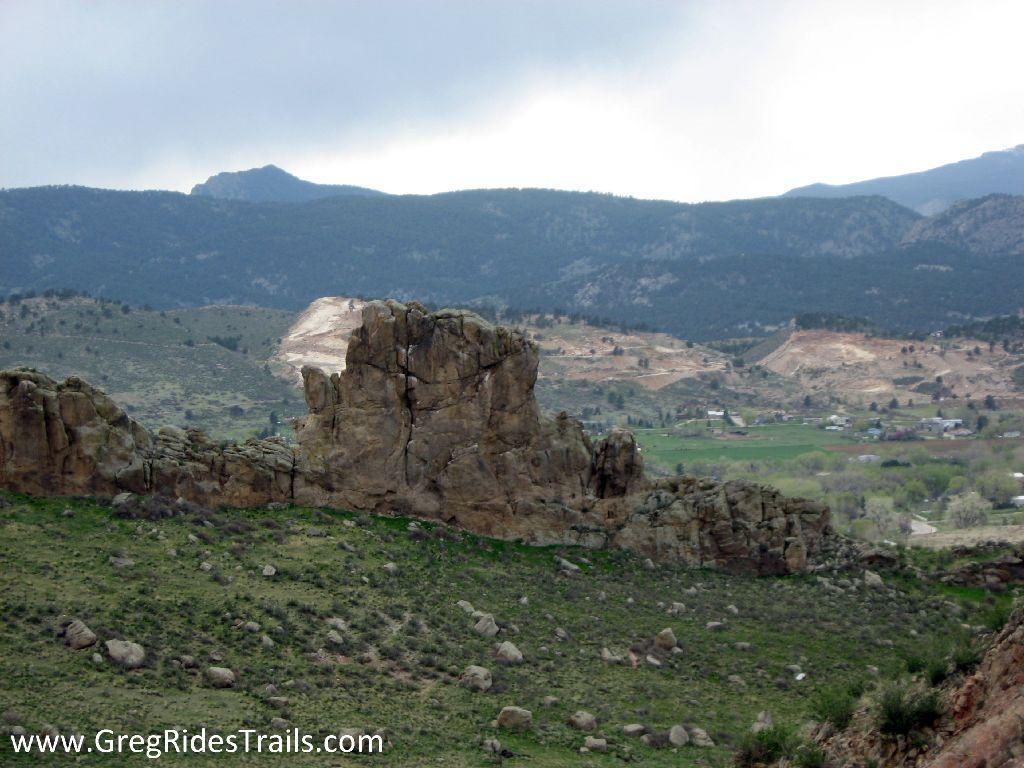 A rugged rock formation stands prominently in the foreground, surrounded by rolling hills and grassy terrain. In the background, a range of mountains stretches under a cloudy sky, with patches of clear blue visible. A small valley can be seen, featuring a mix of tree cover and open fields, hinting at human habitation in the distance. Devil's Backbone mountain bike trail.