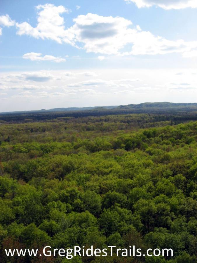 A panoramic view of lush green forests under a partly cloudy sky, showcasing rolling hills in the distance and a vibrant landscape filled with trees during the day. Levis Mounds mountain bike trail.