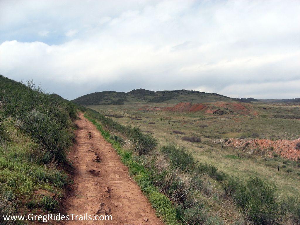 A dirt trail winding through a grassy landscape, flanked by low shrubs and hills in the background under a cloudy sky. Devil's Backbone mountain bike trail.