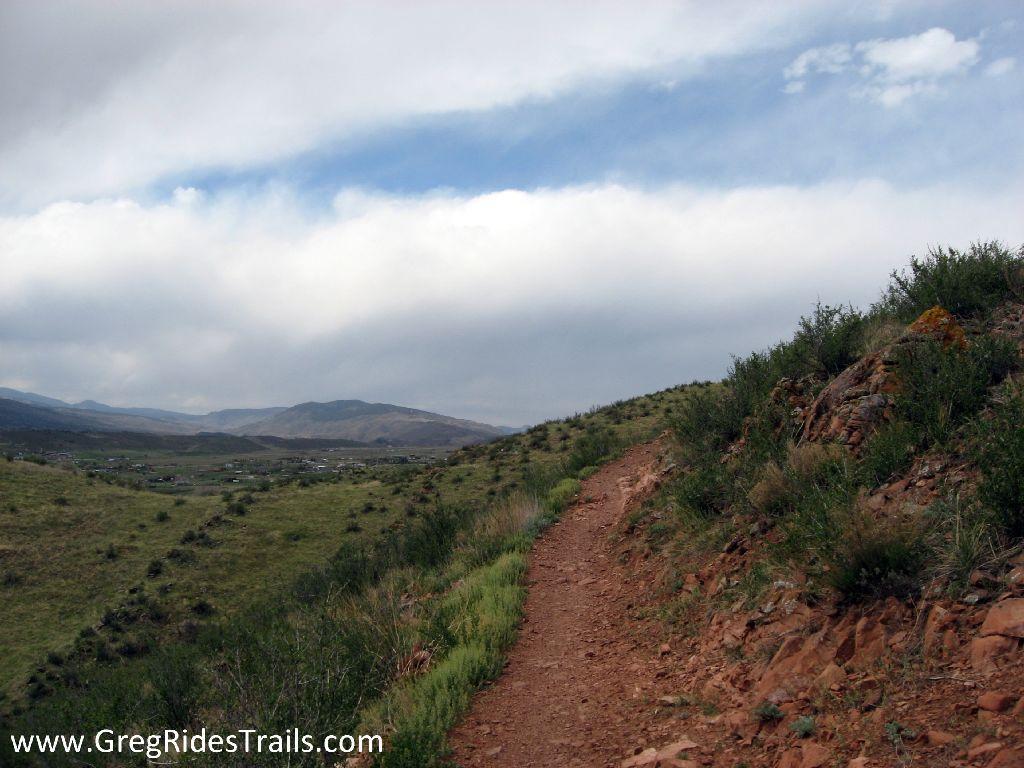 A winding dirt trail surrounded by greenery and rocky terrain, leading through a hilly landscape under a cloudy sky. The view includes distant mountains and a small settlement in the valley below. Devil's Backbone mountain bike trail.