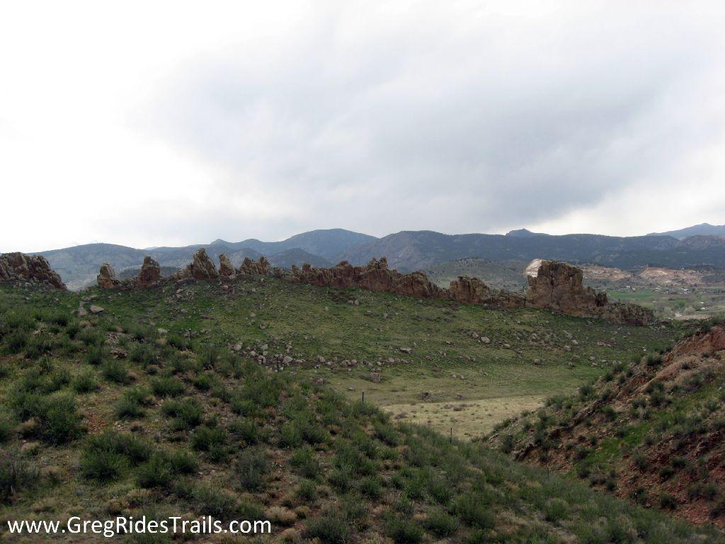 A panoramic view of rolling green hills and rocky formations under a cloudy sky, with distant mountains in the background. The landscape showcases natural vegetation and rugged terrain. Devil's Backbone mountain bike trail.