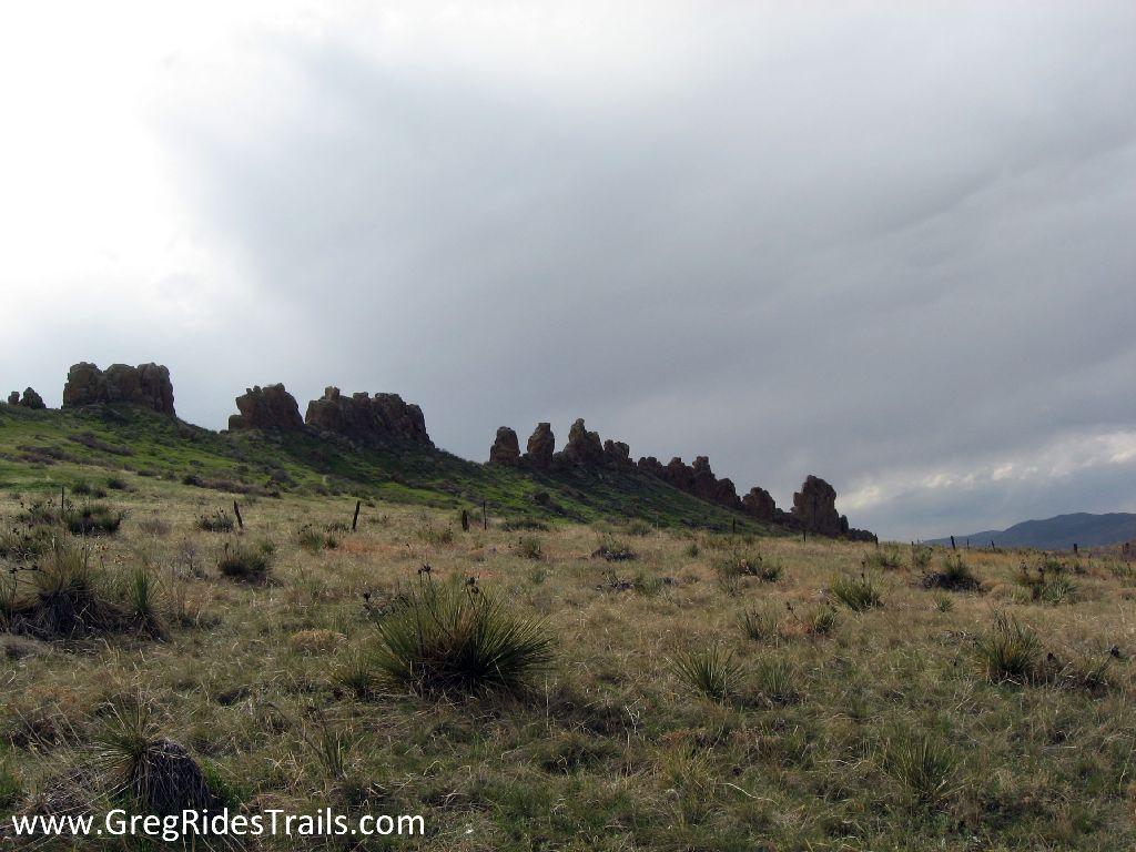 A wide view of rocky formations on a grassy hillside under a cloudy sky, with sparse vegetation in the foreground and a distant mountainous landscape. Devil's Backbone mountain bike trail.