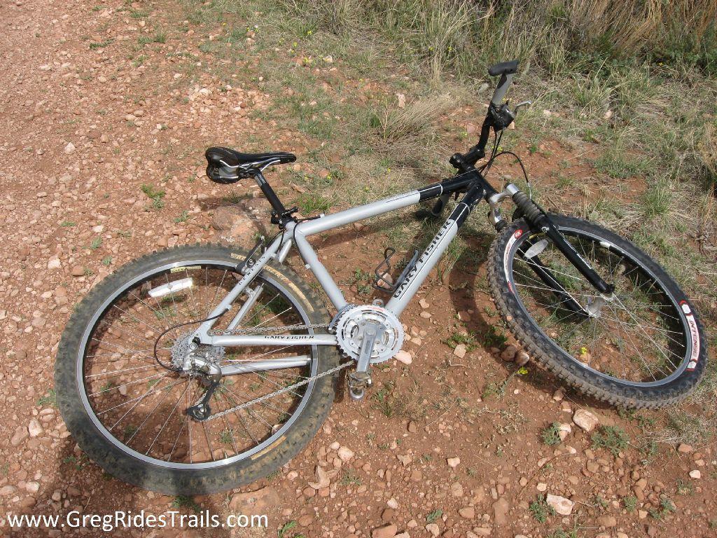 A silver and black mountain bike lying on a dirt trail with gravel and small plants around it. Coyote Ridge mountain bike trail.