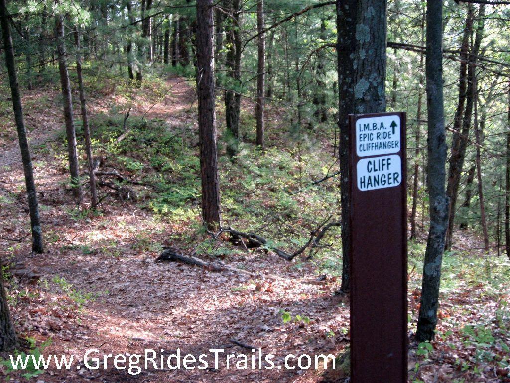 A wooded trail path in a forest, featuring a signpost that reads "I.M.B.A. Epic Ride Cliffhanger" and "Cliff Hanger." The trail is surrounded by trees and covered in fallen leaves, with a slight curve leading into the distance. Levis Mounds mountain bike trail.