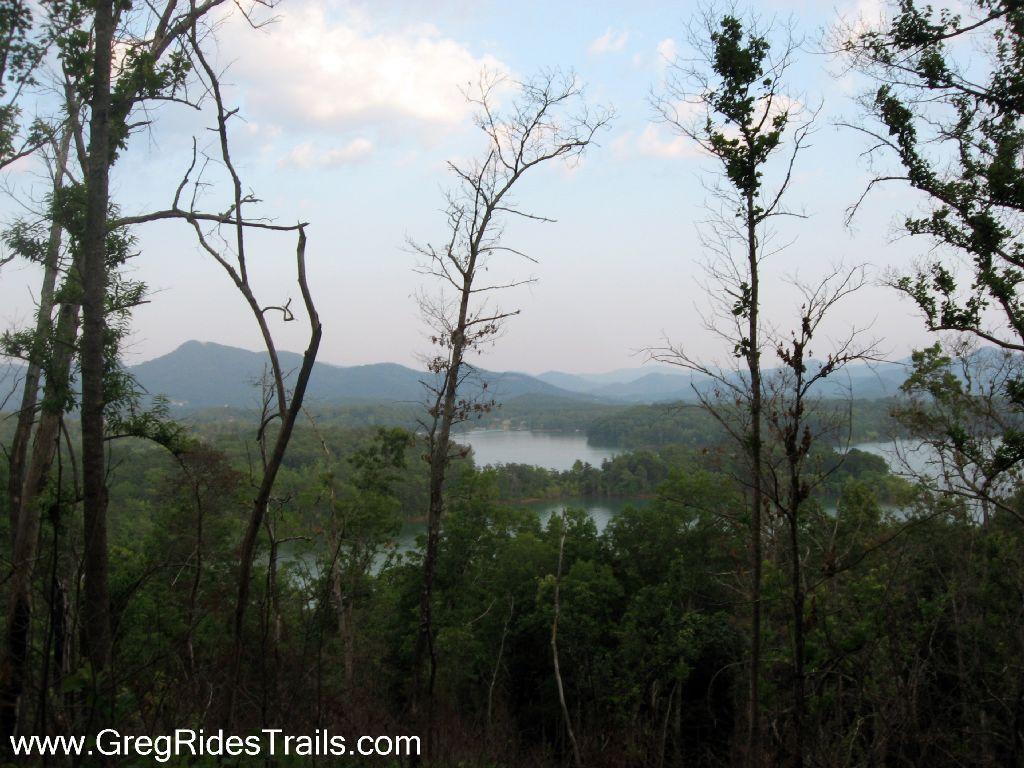 A serene landscape view featuring a calm lake surrounded by lush greenery and distant mountains under a partly cloudy sky. The foreground includes tall, bare trees, adding depth to the scene. Jack Rabbit Trails mountain bike trail.