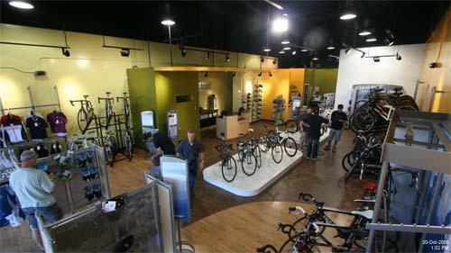 A spacious bicycle shop interior featuring various styles of bicycles and cycling gear. The shop has bright lighting and a modern design, with customers browsing and staff assisting them. Display racks showcase clothing and accessories, while a central area highlights several bicycles.