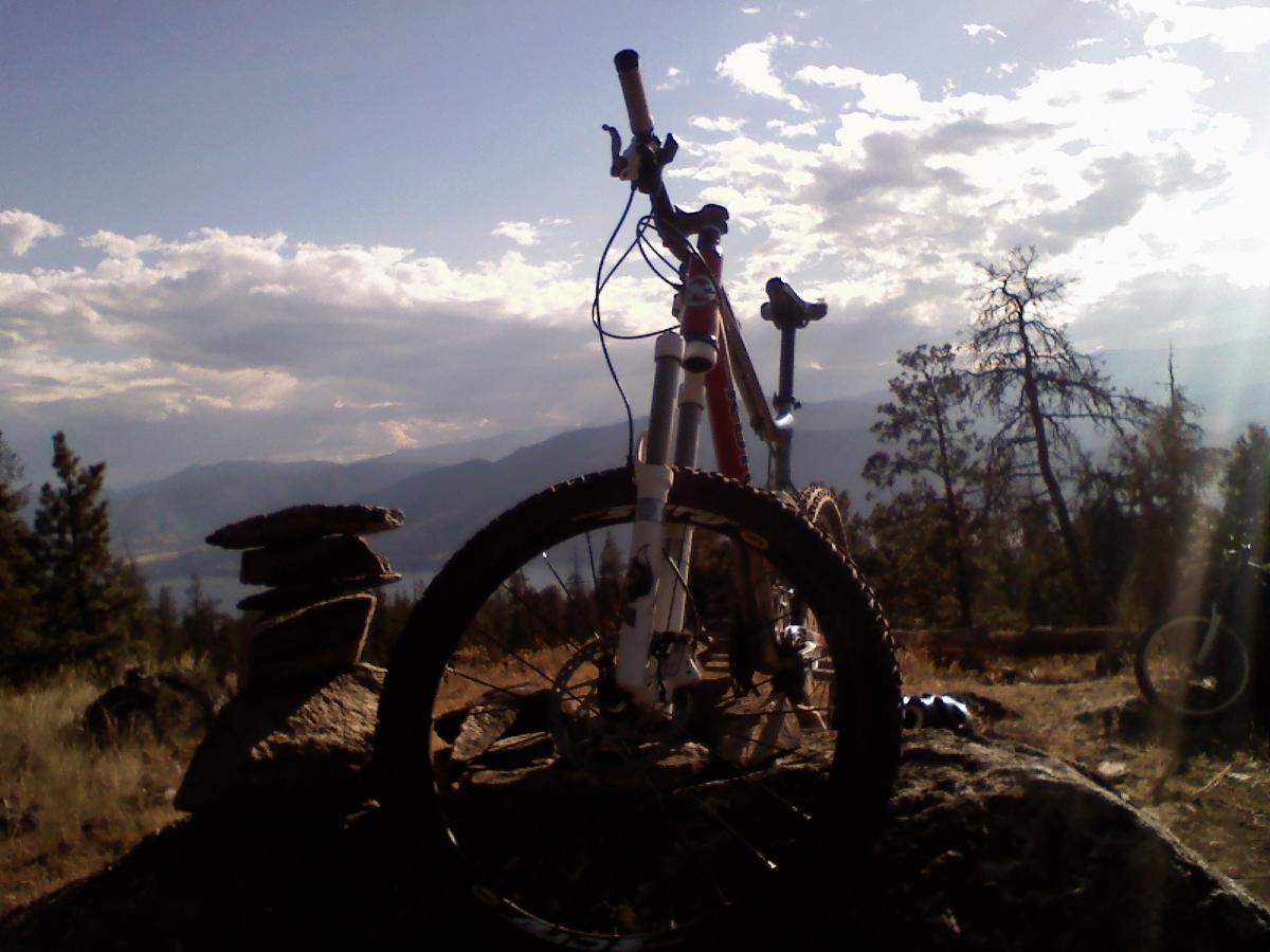 A mountain bike resting on a rock with stacked stones nearby, set against a scenic backdrop of hills, trees, and a cloudy sky. The sun is shining through the clouds, creating a beautiful outdoor atmosphere. Three Blind Mice Trail Network mountain bike trail.