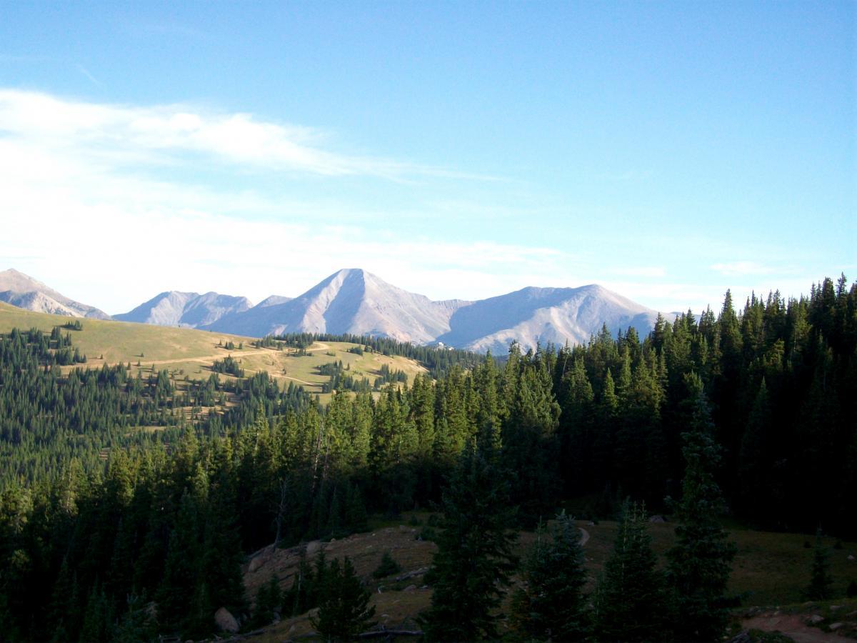 A scenic view of mountainous terrain featuring a mix of rolling hills and evergreen forests under a clear blue sky. The photo showcases several prominent peaks in the distance, highlighting the natural beauty of the landscape. Monarch Crest Trail mountain bike trail.