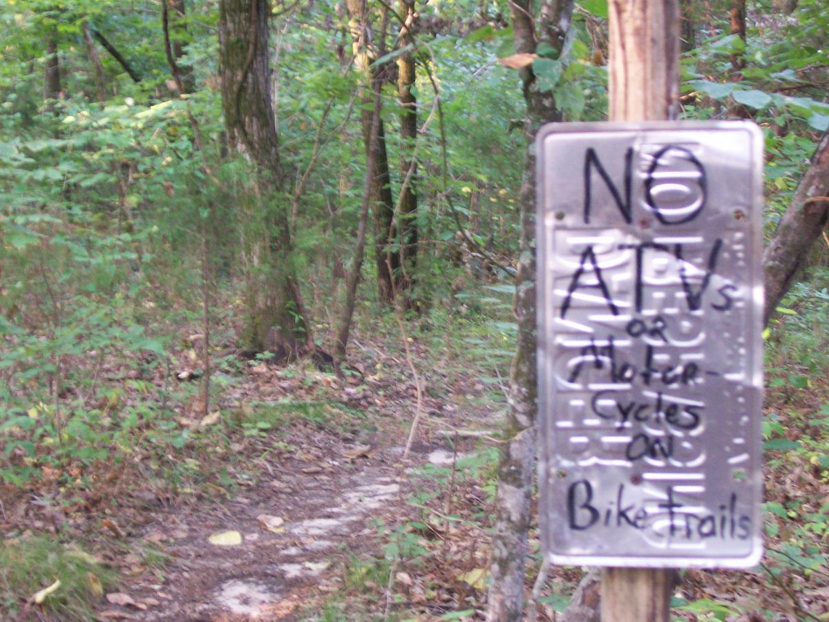 A metal sign on a wooded trail stating "No ATVs or Motorcycles on Bike Trails," with handwritten notes. The background features trees and a dirt path leading into the forest. Research and Technology Park (ERC) mountain bike trail.