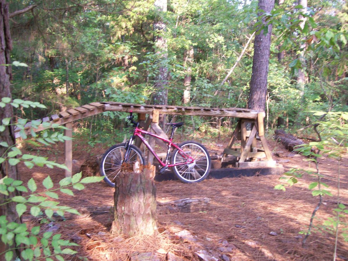 A pink mountain bike is leaning against a wooden structure in a forested area, surrounded by trees and pine needles on the ground. The structure appears to be a ramp or platform, made of wooden planks, set against a natural backdrop of greenery. Research and Technology Park (ERC) mountain bike trail.