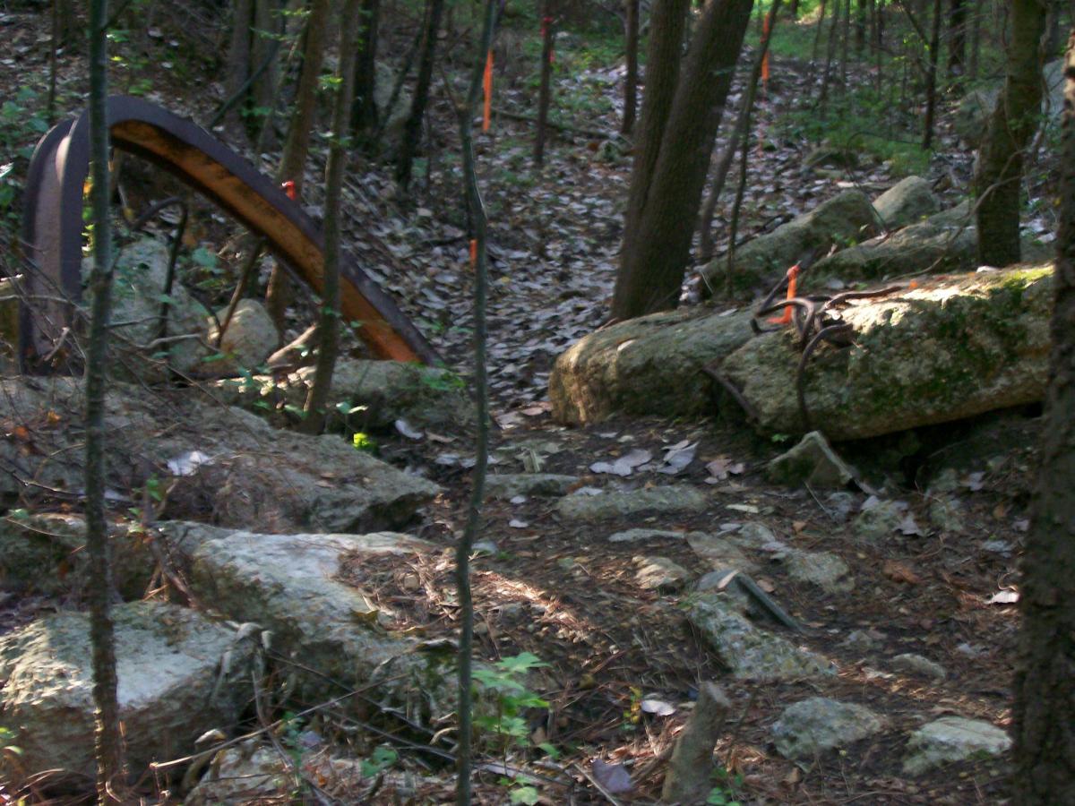 A pathway through a wooded area, featuring large rocks and fallen leaves, with an old metal structure partially visible among the trees. Orange survey tape is marking the surrounding area. Research and Technology Park (ERC) mountain bike trail.
