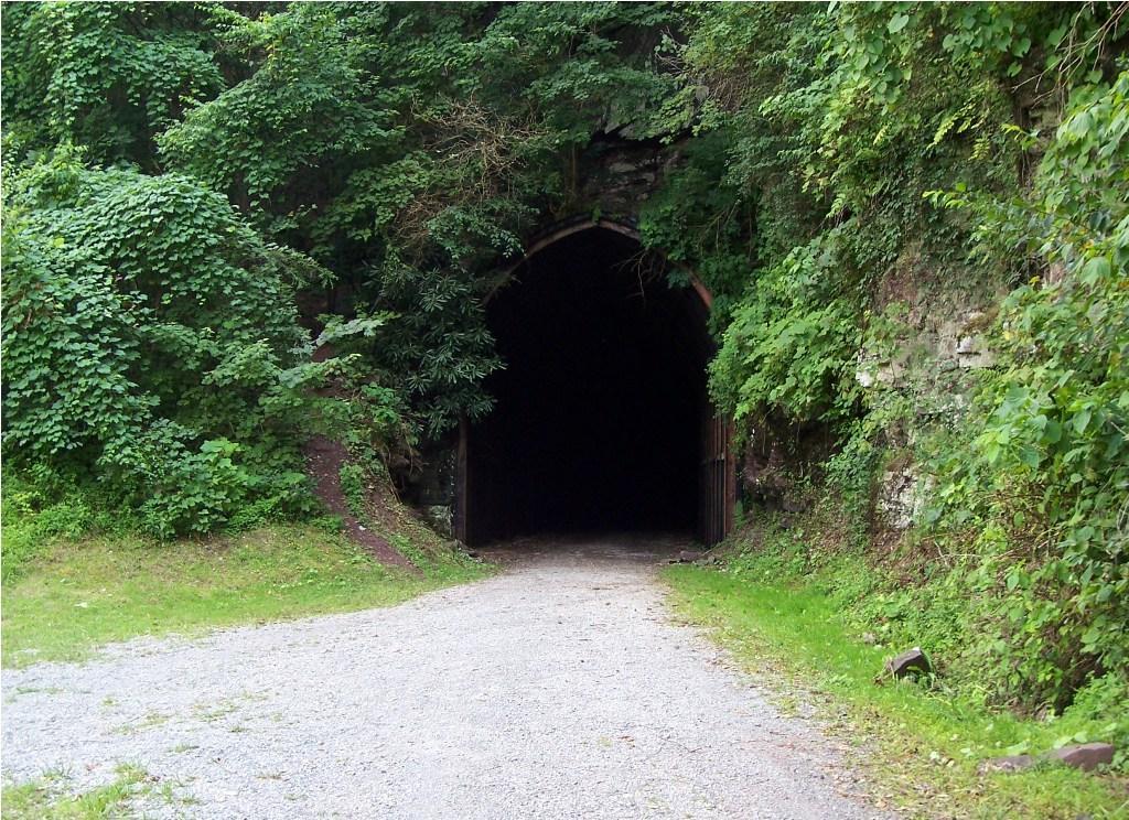 A gravel path leads to a dark tunnel entrance, surrounded by lush green foliage and stone walls. The tunnel appears to be unlit, creating a strong contrast between the bright greenery and the shadowy interior. Greenbrier River Trail mountain bike trail.