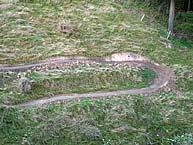 A winding dirt path with a grassy landscape surrounding it, showing curves and turns. Switchback mountain bike trail.