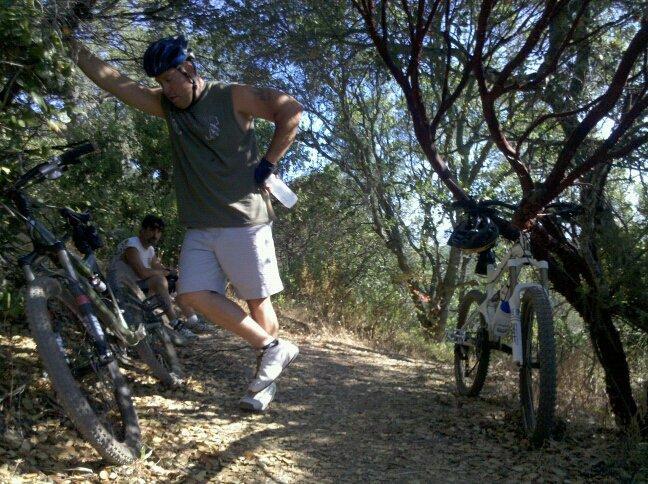 A man in a sleeveless shirt and cycling shorts stretches or adjusts his leg while leaning against a tree on a dirt trail, with mountain bikes parked nearby. Another person sits in the background, surrounded by greenery. The scene is sunny, showcasing a natural setting for outdoor cycling. Rockville Park mountain bike trail.