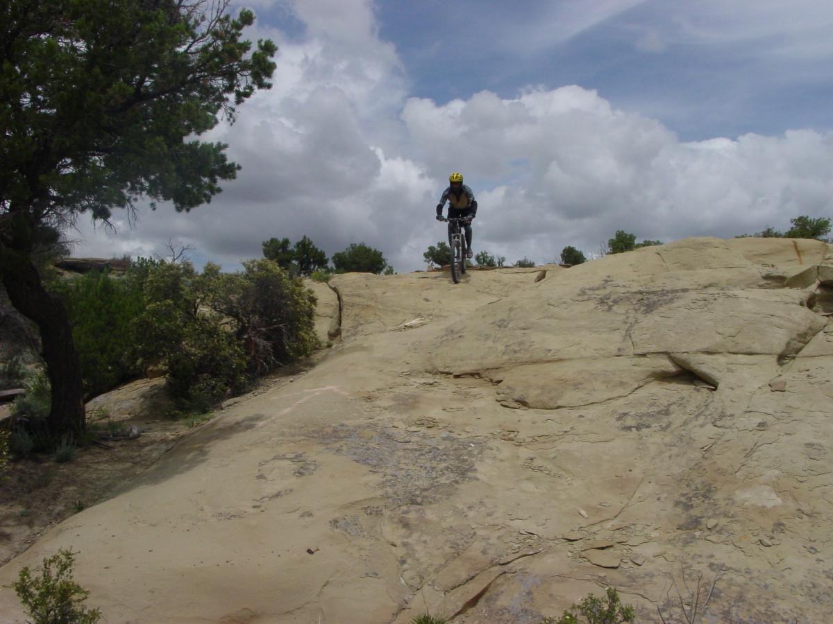 A mountain biker descending a rocky slope under a cloudy sky, surrounded by sparse vegetation and trees. High Desert Trail System mountain bike trail.