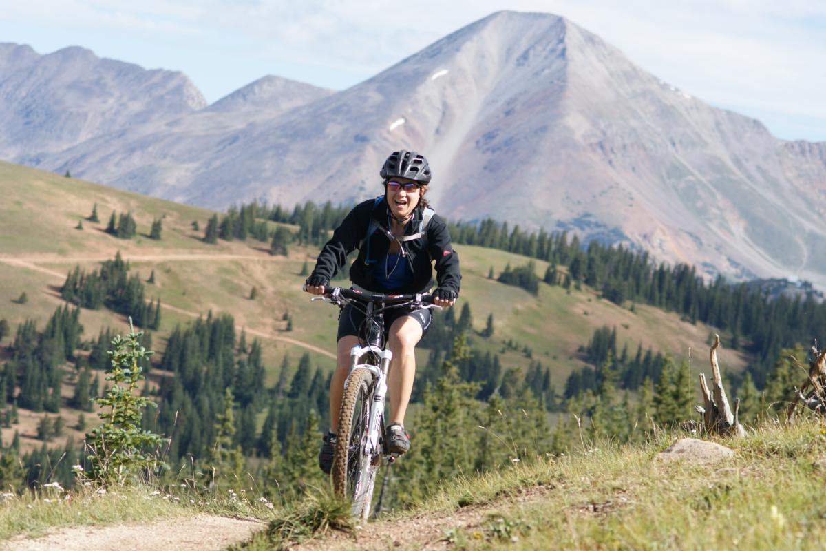 A person riding a mountain bike on a trail in a mountainous landscape, surrounded by green grass and pine trees, with a backdrop of high peaks under a blue sky. The rider is wearing a helmet and appears to be enjoying the ride. Monarch Crest Trail mountain bike trail.