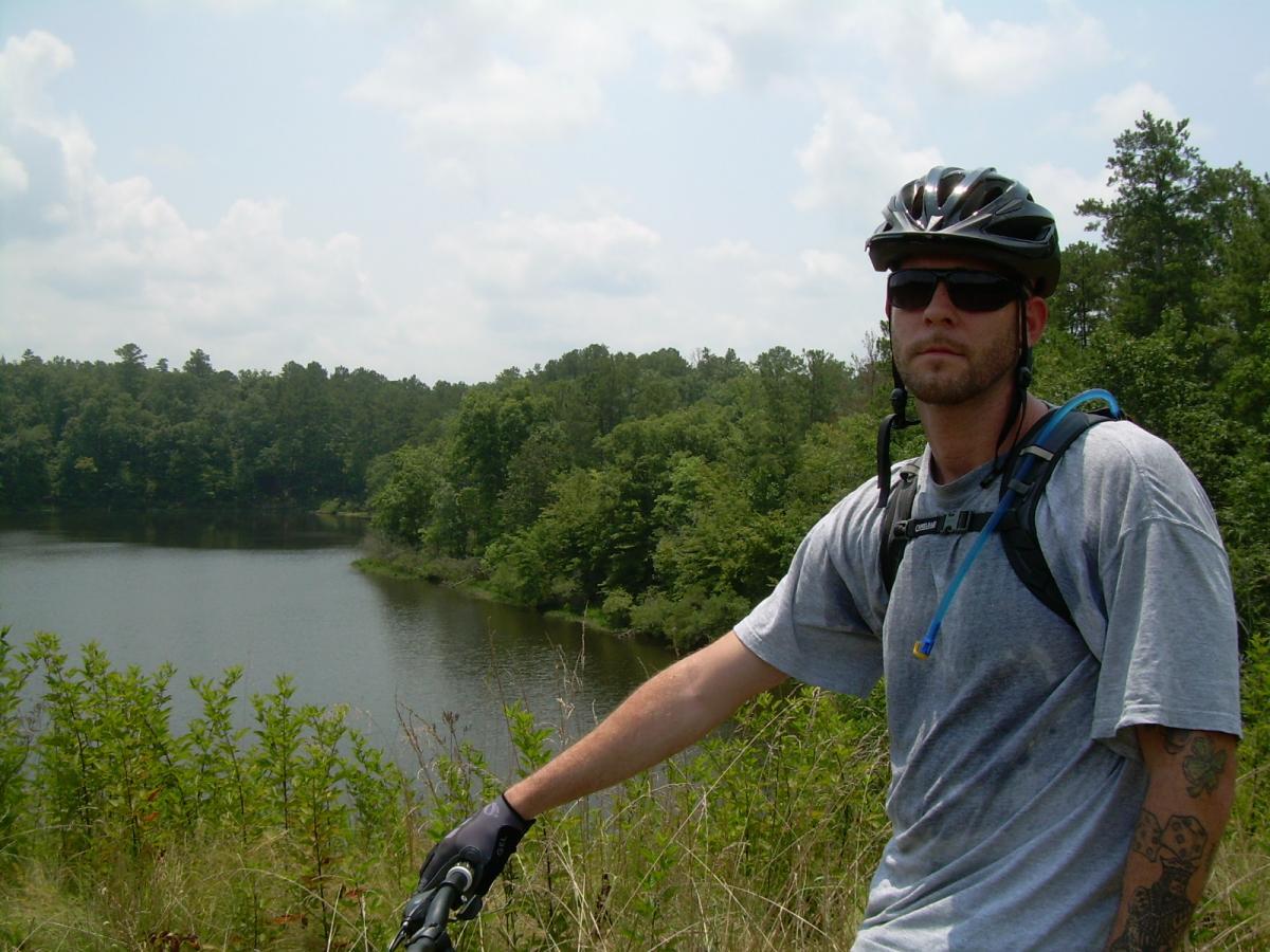 A person wearing a helmet and sunglasses stands beside a mountain bike, overlooking a calm lake surrounded by lush green trees under a partly cloudy sky. The individual is dressed in a gray t-shirt and has a hydration pack. Sylaward mountain bike trail.