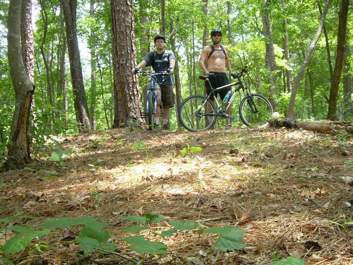 Two men standing with their mountain bikes in a wooded area. One is wearing a helmet and a t-shirt, while the other is shirtless, with sunglasses and shorts. The ground is covered in pine needles and small plants, surrounded by tall trees and greenery. Sylaward mountain bike trail.