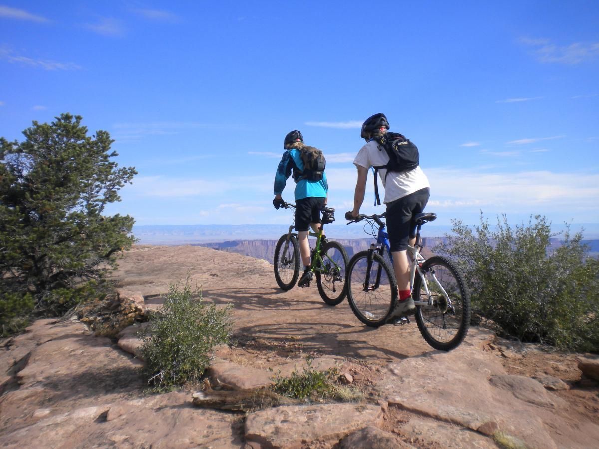 Two mountain bikers standing on a rocky ledge, overlooking a vast landscape with a clear blue sky. One rider is wearing a blue jacket and the other is in a white shirt, both equipped with helmets and backpacks, enjoying the scenic view. UPS And LPS mountain bike trail.