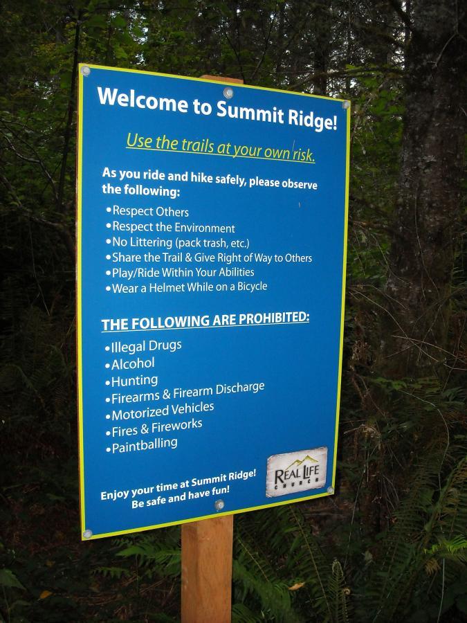 A blue informational sign at Summit Ridge detailing safety guidelines for hikers and bikers. The sign emphasizes the importance of respecting others and the environment, sharing the trail, riding within one's abilities, and wearing helmets on bicycles. It also lists prohibited activities, including the use of illegal drugs, alcohol, hunting, firearms, motorized vehicles, fires, and paintballing. The sign concludes with a message encouraging visitors to enjoy their time safely at Summit Ridge. Lake Sawyer mountain bike trail.