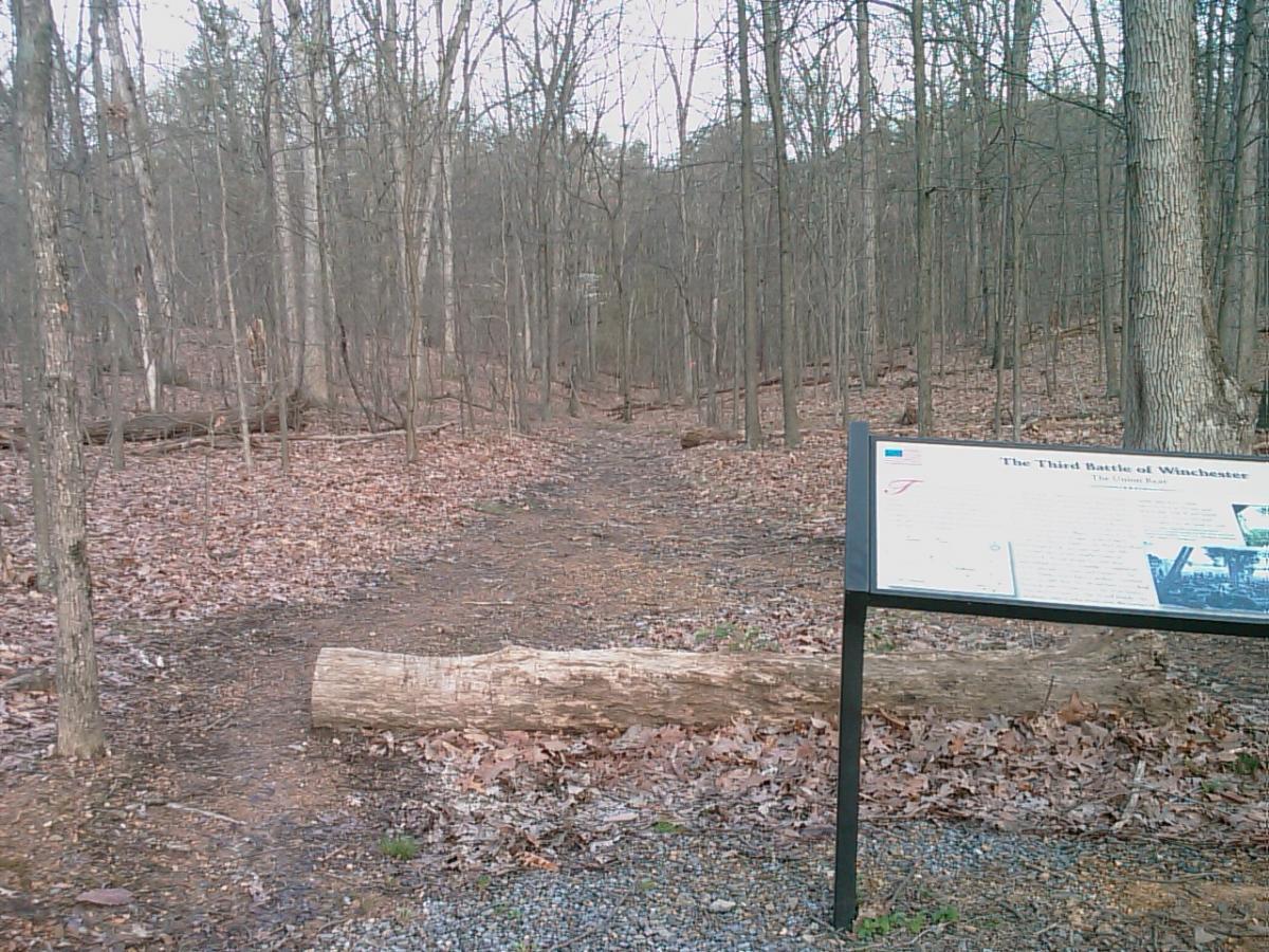 Sign marking the site of the Third Battle of Winchester, positioned along a forested path. The area features bare trees, fallen leaves, and a log lying across the trail, creating a natural, historical setting. 3rd Battle Of Winchester Trail mountain bike trail.