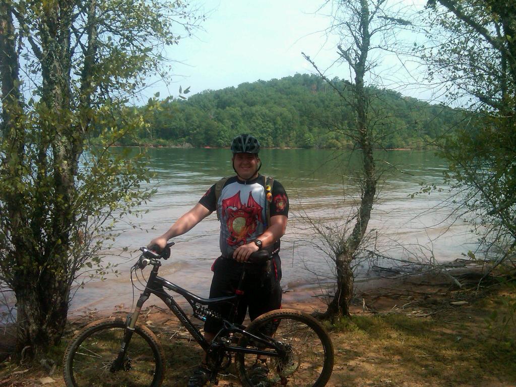 A person in a black helmet and a colorful cycling jersey stands beside a mountain bike near a calm body of water, with trees and a green hillside in the background on a sunny day. Jack Rabbit Trails mountain bike trail.