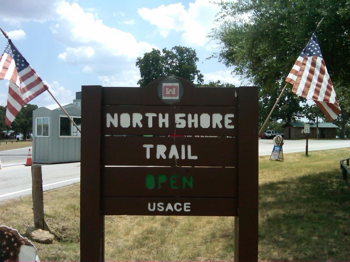 Sign indicating the North Shore Trail is open, with two American flags on either side. The sign is wooden and has "NORTH SHORE TRAIL" written prominently, along with "OPEN" and "USACE" at the bottom. In the background, a small building and grassy area are visible under a partly cloudy sky. Northshore Trail mountain bike trail.
