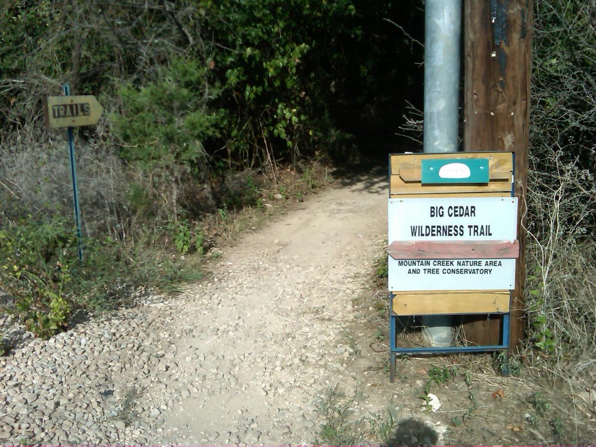 A trail sign marking the entrance to the Big Cedar Wilderness Trail, located in the Mountain Creek Nature Area and Tree Conservatory. The path is surrounded by dense vegetation and gravel, with another sign indicating nearby trails. Big Cedar Wilderness Trails mountain bike trail.