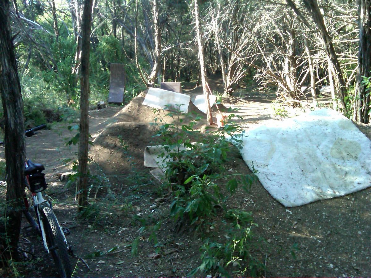 A wooded area featuring a dirt bike ramp and jump. In the foreground, a bike rests against a tree. The background includes several dirt mounds and ramps made of wood, surrounded by dense foliage and trees. The scene is sunny and natural, perfect for biking activities. Big Cedar Wilderness Trails mountain bike trail.
