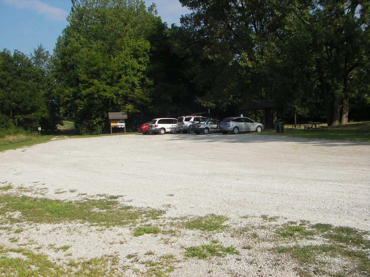 A gravel parking lot surrounded by trees, with several parked cars and a wooden information sign at the entrance to a trail. Clear blue sky above enhances the natural setting. Kickapoo mountain bike trail.