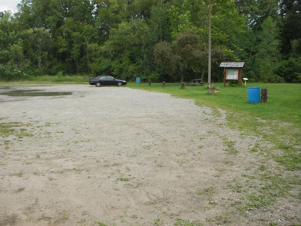 A gravel parking area surrounded by greenery, featuring a black car parked on the right. In the background, there is a signboard and a few wooden picnic tables, with a blue barrel nearby. The scene is tranquil, with trees lining the area. Outback Trail at Imagination Glenn mountain bike trail.