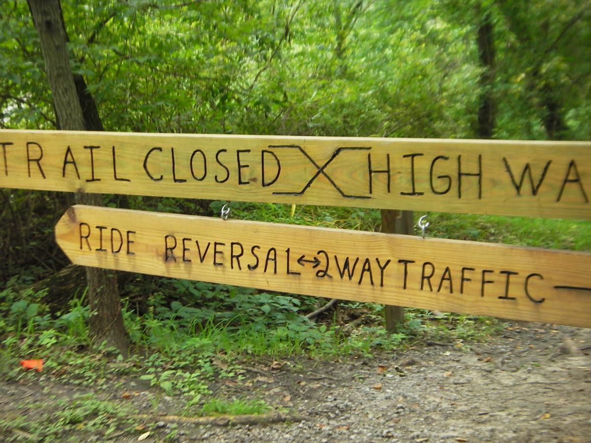 Wooden trail sign indicating "Trail Closed" overlaid with an X and another sign below stating "Ride Reversal ↔ 2 Way Traffic," set against a lush green background of trees and foliage. Outback Trail at Imagination Glenn mountain bike trail.