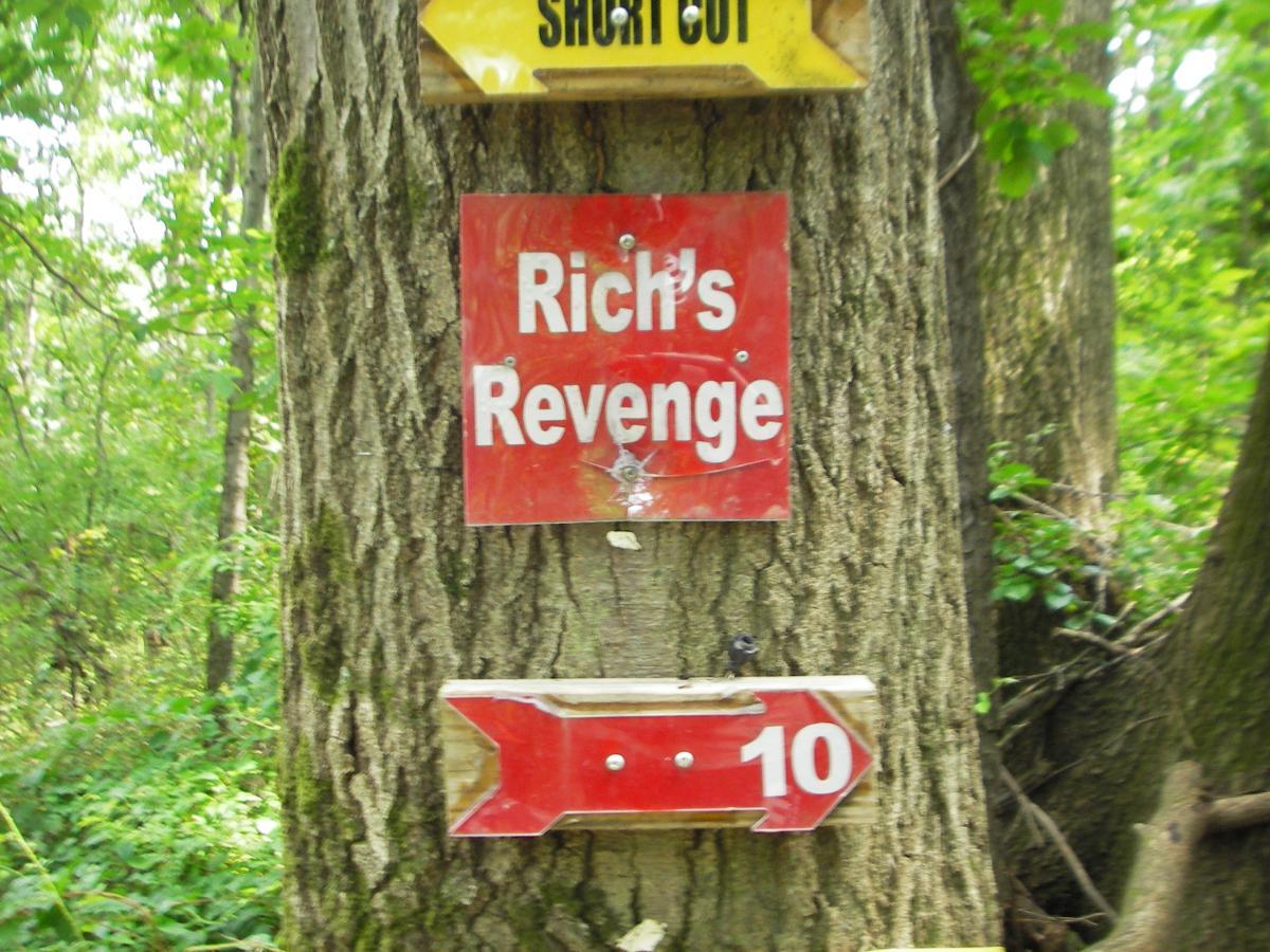 A red sign mounted on a tree in a wooded area, displaying the text "Rich's Revenge" in white letters. Below it, there is an arrow pointing to the right, accompanied by the number "10." Other directional signs can be seen above, indicating a shortcut. The background features lush green foliage. Outback Trail at Imagination Glenn mountain bike trail.