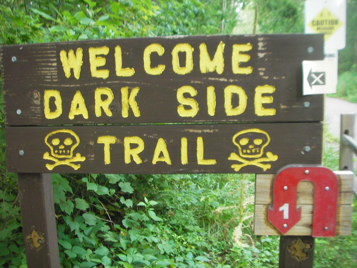 A wooden sign with yellow lettering that reads "Welcome Dark Side Trail," featuring skull and crossbones illustrations. Below the main sign, there is a red arrow pointing downwards with the number "1." The sign is surrounded by lush green foliage, indicating a wooded area. Outback Trail at Imagination Glenn mountain bike trail.