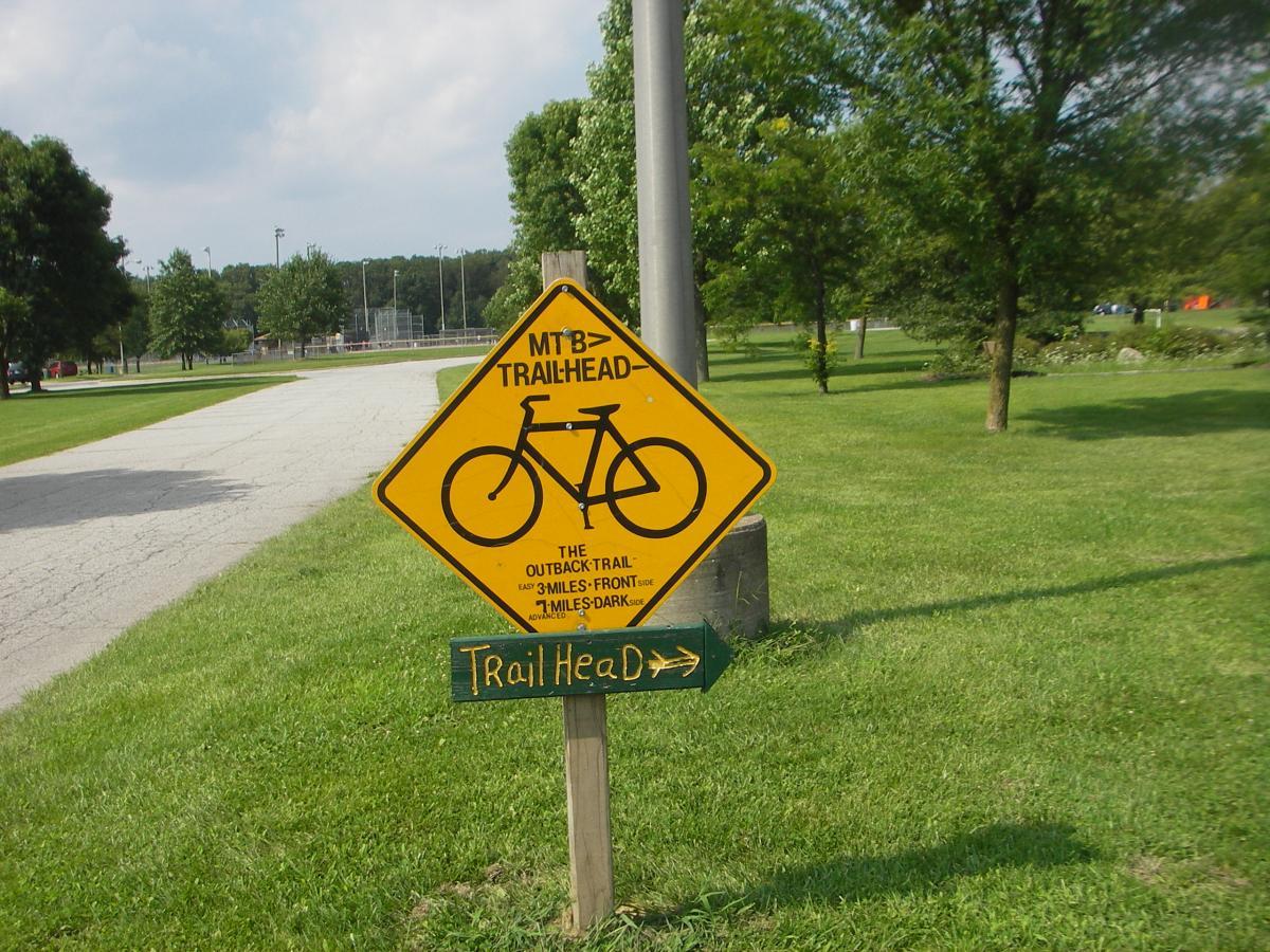A yellow trailhead sign with a bicycle graphic, indicating a mountain biking trail. The sign reads "MTB TRAILHEAD" and provides directions to the "Outback Trail," which is located three miles ahead from the current position. A green arrow points to the right, suggesting the direction to the trail. The background features green grass and trees, along with a paved path leading into a park area. Outback Trail at Imagination Glenn mountain bike trail.