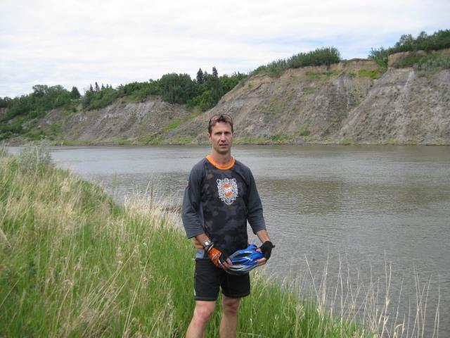 A person standing by a riverbank surrounded by tall grass and a steep, grassy cliff in the background. The individual is wearing a long-sleeve shirt with a graphic design, shorts, and gloves, holding a bicycle helmet. The scene is set on a cloudy day, with trees visible on the cliff above. Fort Saskatchewan Hidden Gold mountain bike trail.