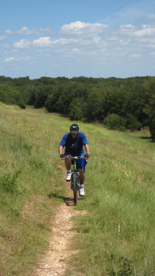A person mountain biking on a dirt trail through a grassy field, surrounded by trees under a partly cloudy sky. The individual is wearing a blue shirt, shorts, and a helmet, focusing on the path ahead. Lake Bryan mountain bike trail.