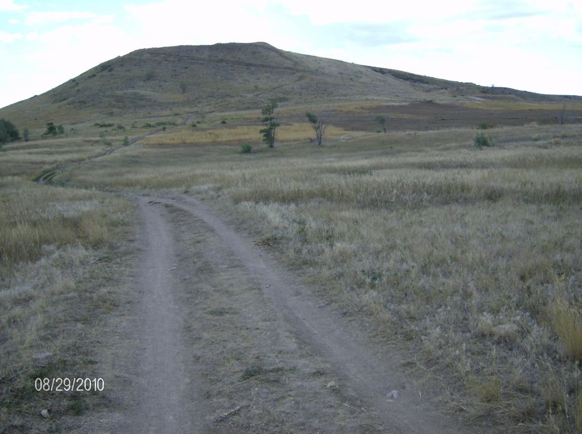 A dirt path leads towards a small, grassy hill in a wide-open landscape. The surrounding area features dry grass and sparse trees, with patches of golden-yellow vegetation evident in the foreground and background. The sky is overcast, adding a muted tone to the scene. Bear Creek Lake Park mountain bike trail.