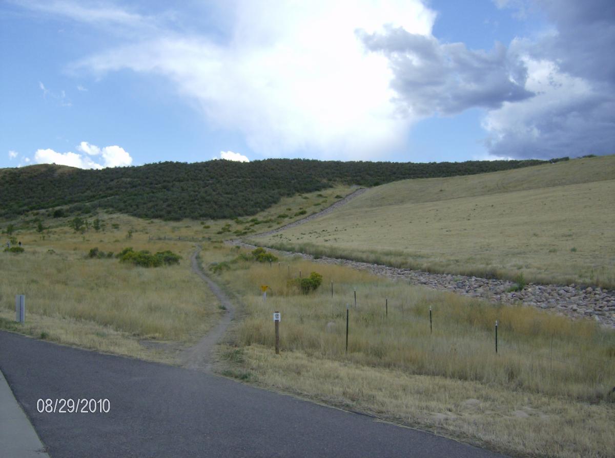 A scenic landscape featuring a grassy hill with patches of shrubs and a winding dirt path leading towards the higher terrain. The foreground includes a paved road, with a date displayed in the corner. The sky is partly cloudy, adding to the natural beauty of the setting. Bear Creek Lake Park mountain bike trail.