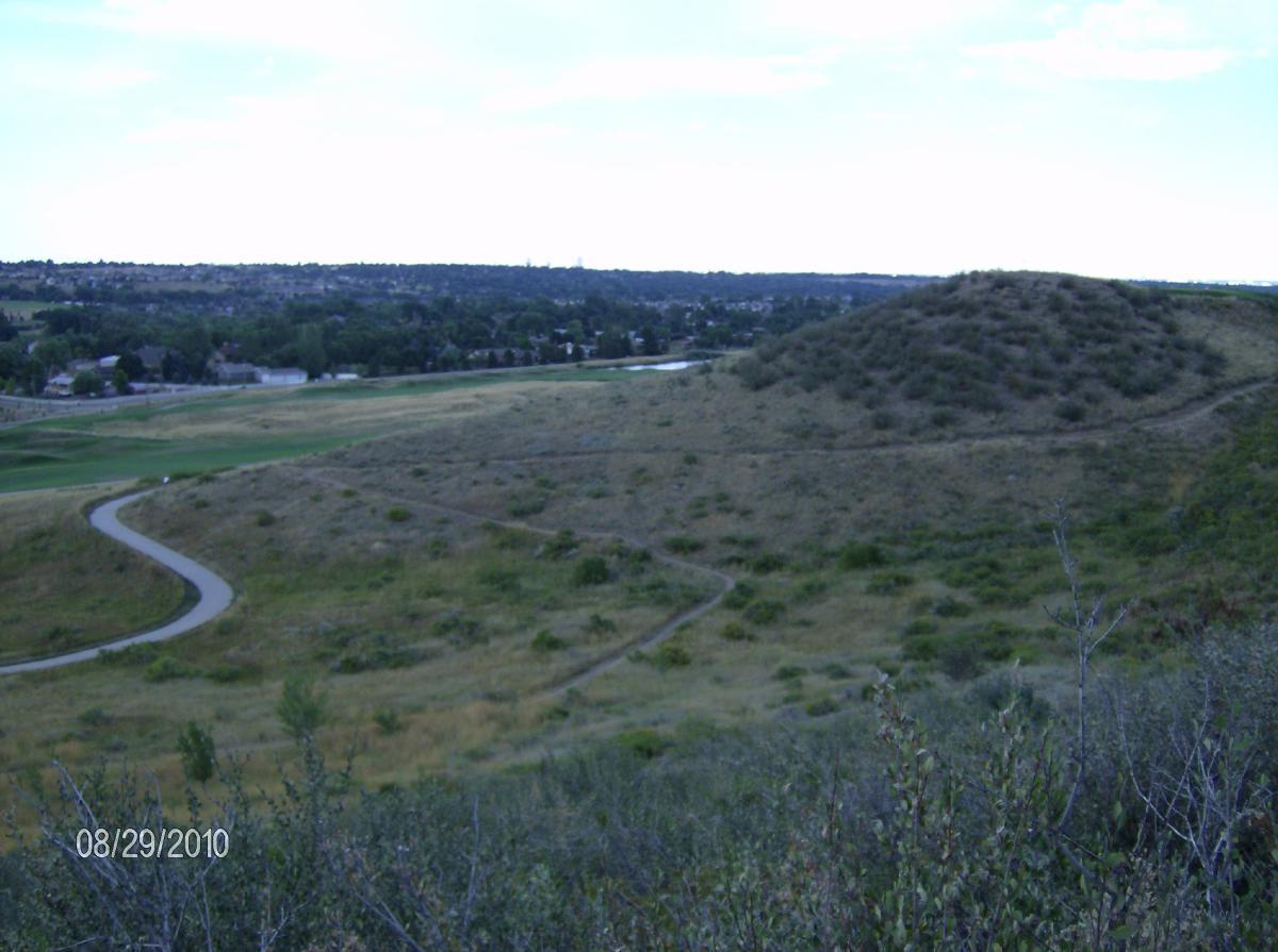 A scenic view of rolling hills and a winding pathway in a grassy landscape, with residential structures in the distance under a cloudy sky. Bear Creek Lake Park mountain bike trail.