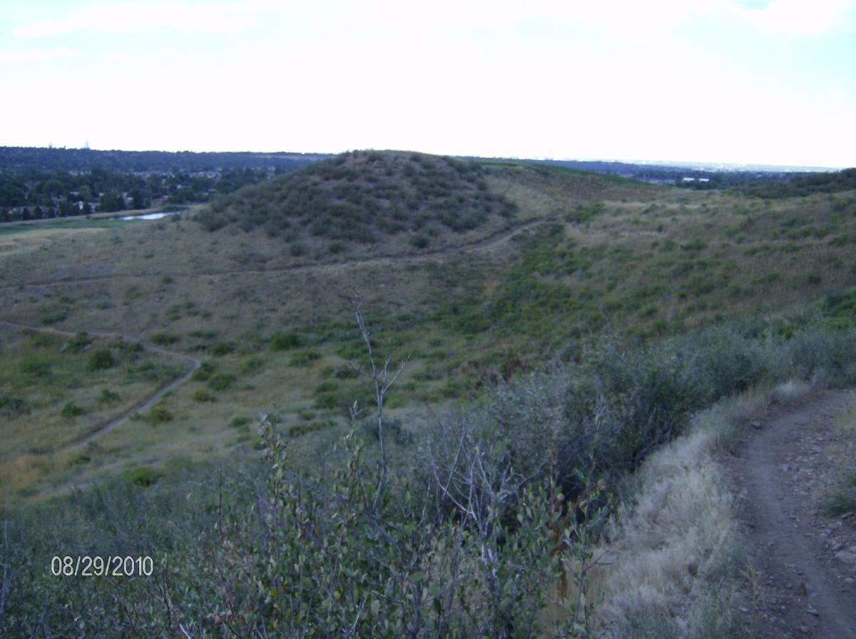A scenic view of rolling hills covered in greenery, with a dirt path winding through the landscape. In the distance, a small body of water is visible, and the skyline of a city can be seen at the horizon under a mostly cloudy sky. The foreground features sparse shrubs and grasses. Bear Creek Lake Park mountain bike trail.