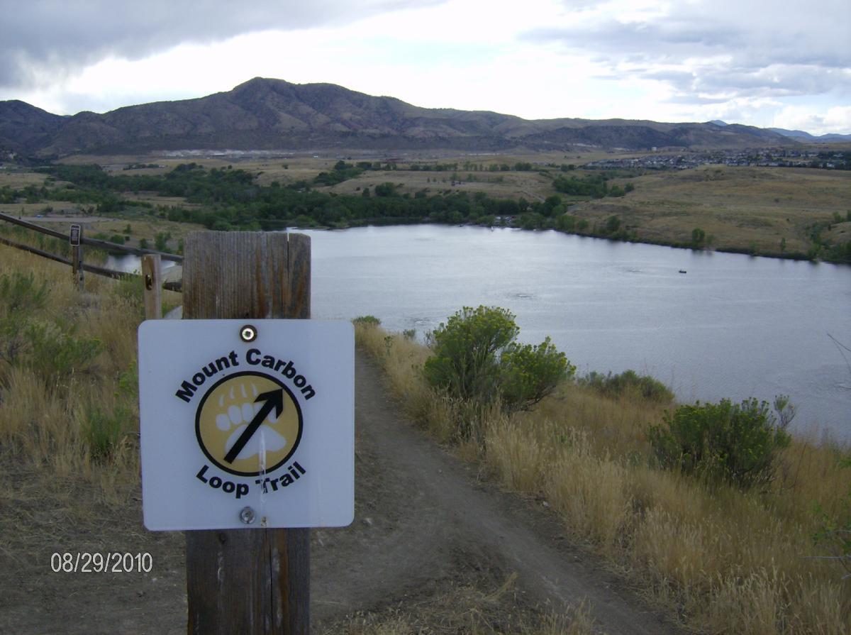 A trail sign for the Mount Carbon Loop Trail points upward, with a scenic view of a lake and rolling hills in the background. The landscape features dry grass and patches of greenery under a cloudy sky. Bear Creek Lake Park mountain bike trail.