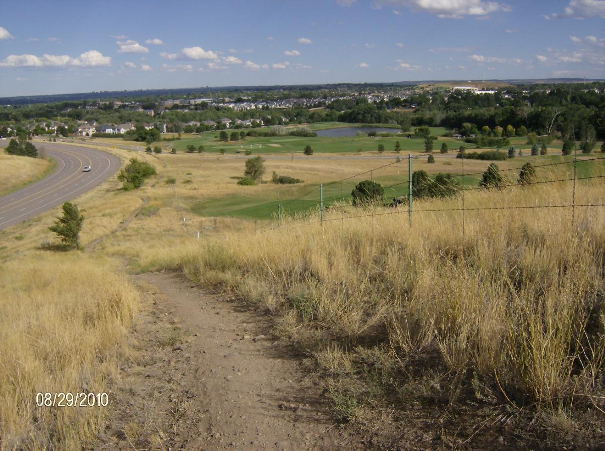 A view from a hillside overlooking a winding road, grassy fields, and a small pond. In the distance, houses can be seen along with areas of trees and a golf course, under a clear blue sky with a few scattered clouds. Bear Creek Lake Park mountain bike trail.